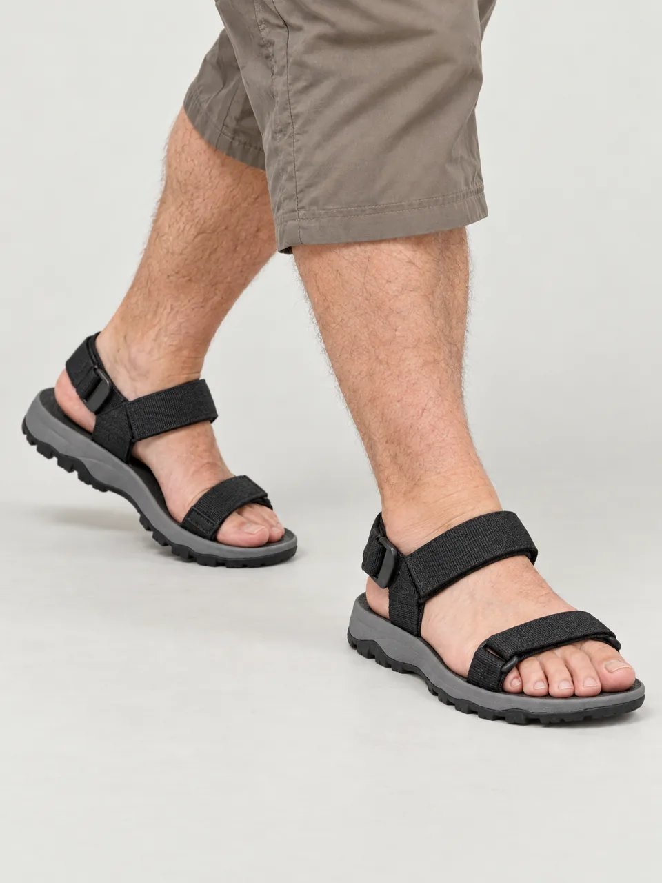 Man in his 50s, white, heavyset and bearded, stepping across a shallow rocky stream in shorts, framed from mid-calf down wearing the Scree Line Men's Tuff Trail Sandal in Black