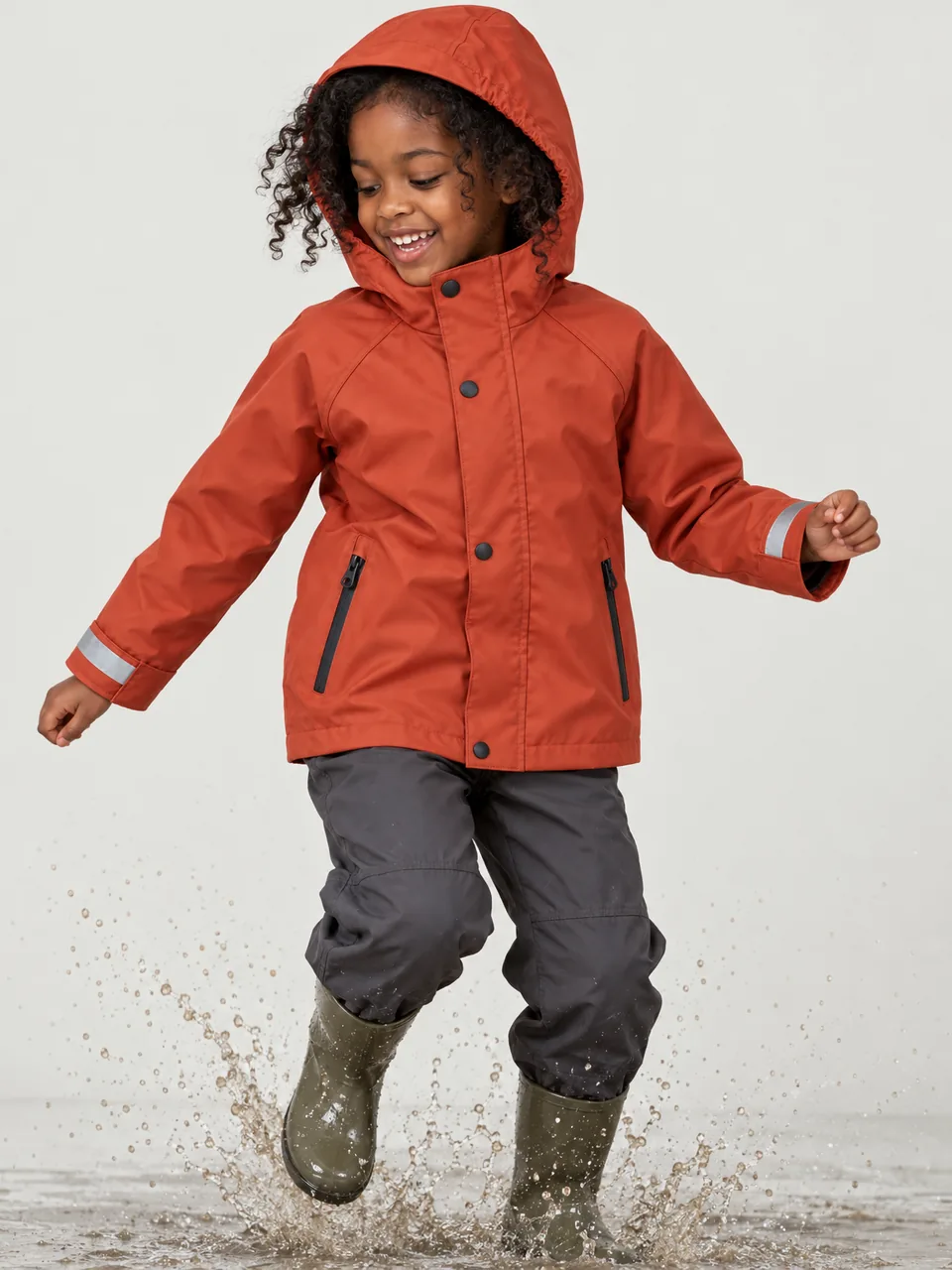 Girl about 7, Black British, mid-jump into a puddle with the hood up, wearing walking trousers and wellies wearing the Hale Kids' Hogg 2.5L Waterproof Jacket in Ember