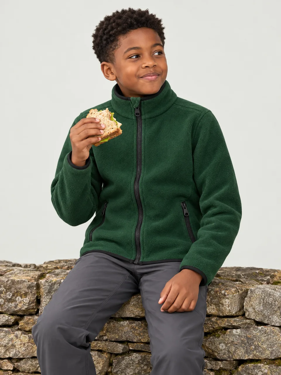 Boy about 11, Black British, sitting on a dry-stone wall eating a sandwich, fleece zipped up wearing the Hale Kids' Clough Fleece Jacket in Holloway
