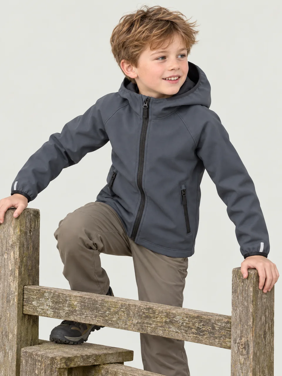 Boy about 8, white, climbing over a wooden stile on a breezy hillside, side-front view wearing the Hale Kids' Brow Softshell Jacket in Slate