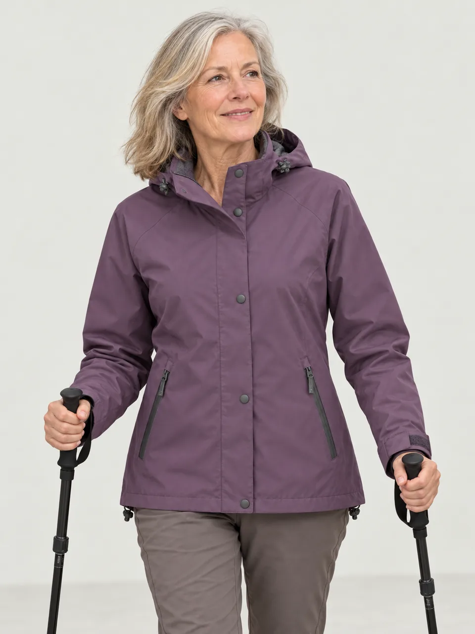 White woman in her 60s, walking beside a lake with walking poles, hood down wearing the Hale Women's Laithe 2L Waterproof Jacket in Heather