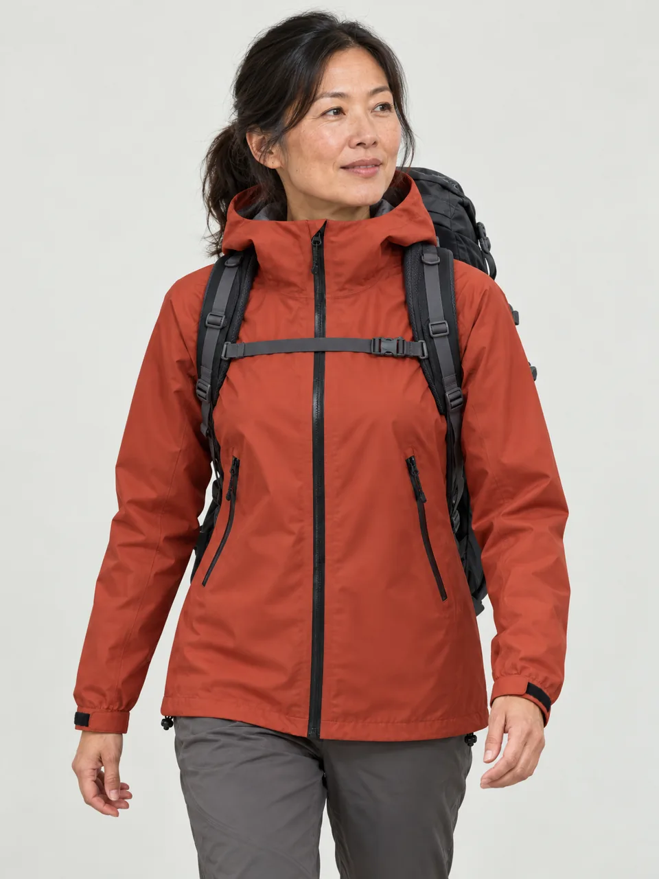 East Asian woman in her 40s, walking a grassy ridge with a 45-litre pack, hood down wearing the Swift Women's Stint 230 3L Waterproof Jacket in Ember