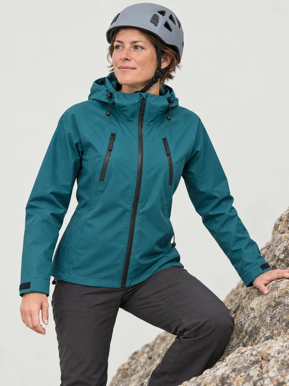 White woman in her 30s, short hair, scrambling on dry rock wearing a helmet, hood down wearing the Cairnhope Women's Smirr 3L Mountain Jacket in Teal