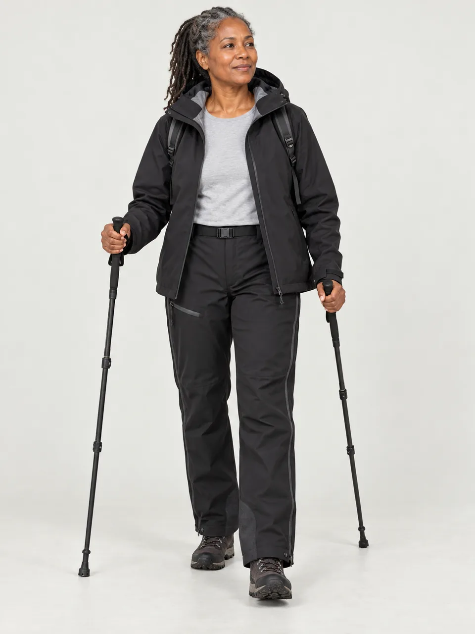 Black woman in her 50s, walking a wet hill path with poles, jacket and overtrousers on wearing the Hale Women's Brackwater 3L Waterproof Trousers in Soot