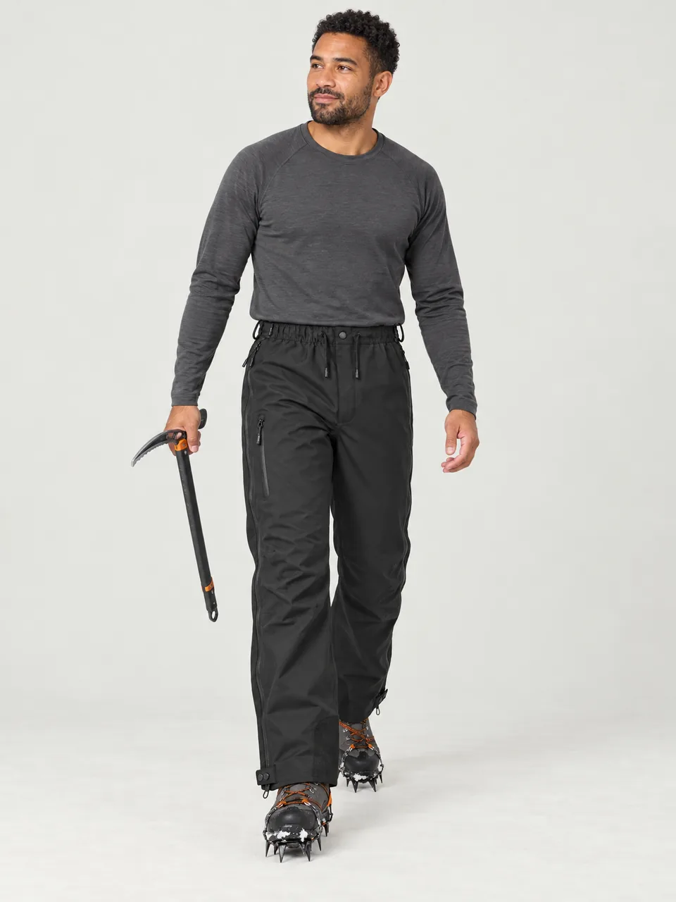 Mixed-race man in his 30s, walking across snow with an ice axe, overtrousers over boots and crampons wearing the Cairnhope Men's Thole 3L Overtrousers in Black