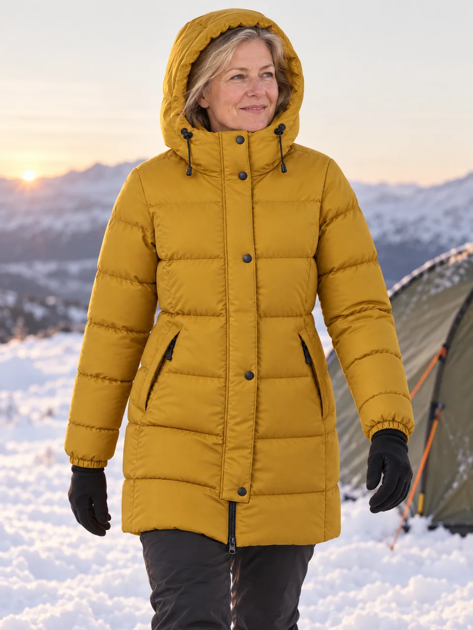 White woman in her 50s, walking through snow beside a tent at dawn, hood up wearing the Northwold Women's Frore 650 Down Parka in Mustard