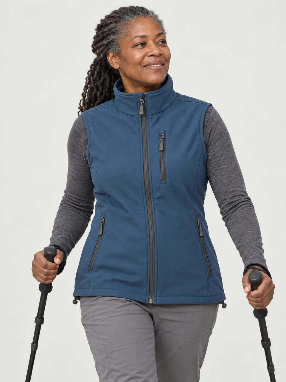 Black woman in her 50s, walking fast on a hill track with poles, gilet over a long-sleeve base layer wearing the Hale Women's Coomb Softshell Gilet in Tarn
