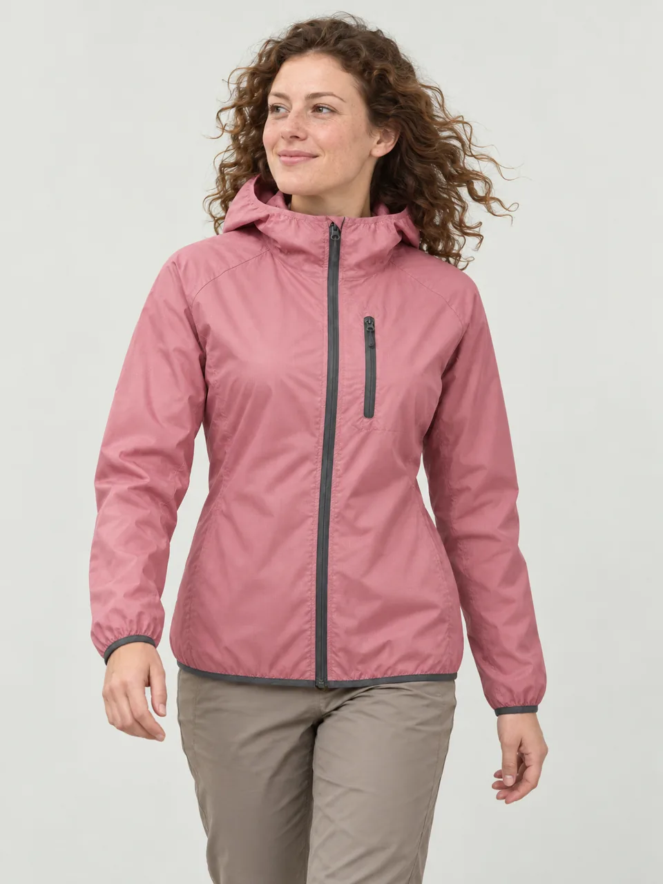 White woman in her 20s, curly hair, walking a clifftop path in a stiff breeze wearing the Hale Women's Sett Windproof Jacket in Thrift