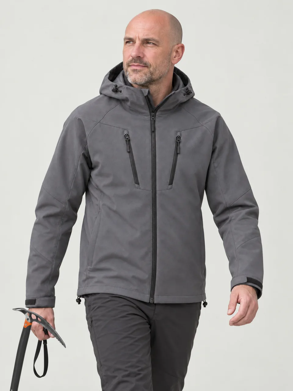 White man in his 40s, shaven head, walking across a snowy plateau with an ice axe in hand, hood down wearing the Cairnhope Men's Haar Softshell Jacket in Storm Grey