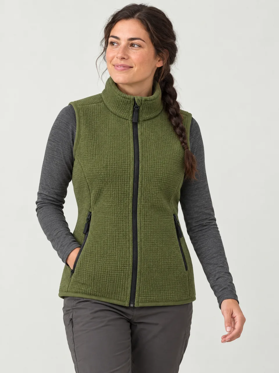 Woman in her 20s with a long dark plait, walking a grassy ridge in a long-sleeve top and the gilet wearing the Hale Women's Sneck Fleece Gilet in Moss