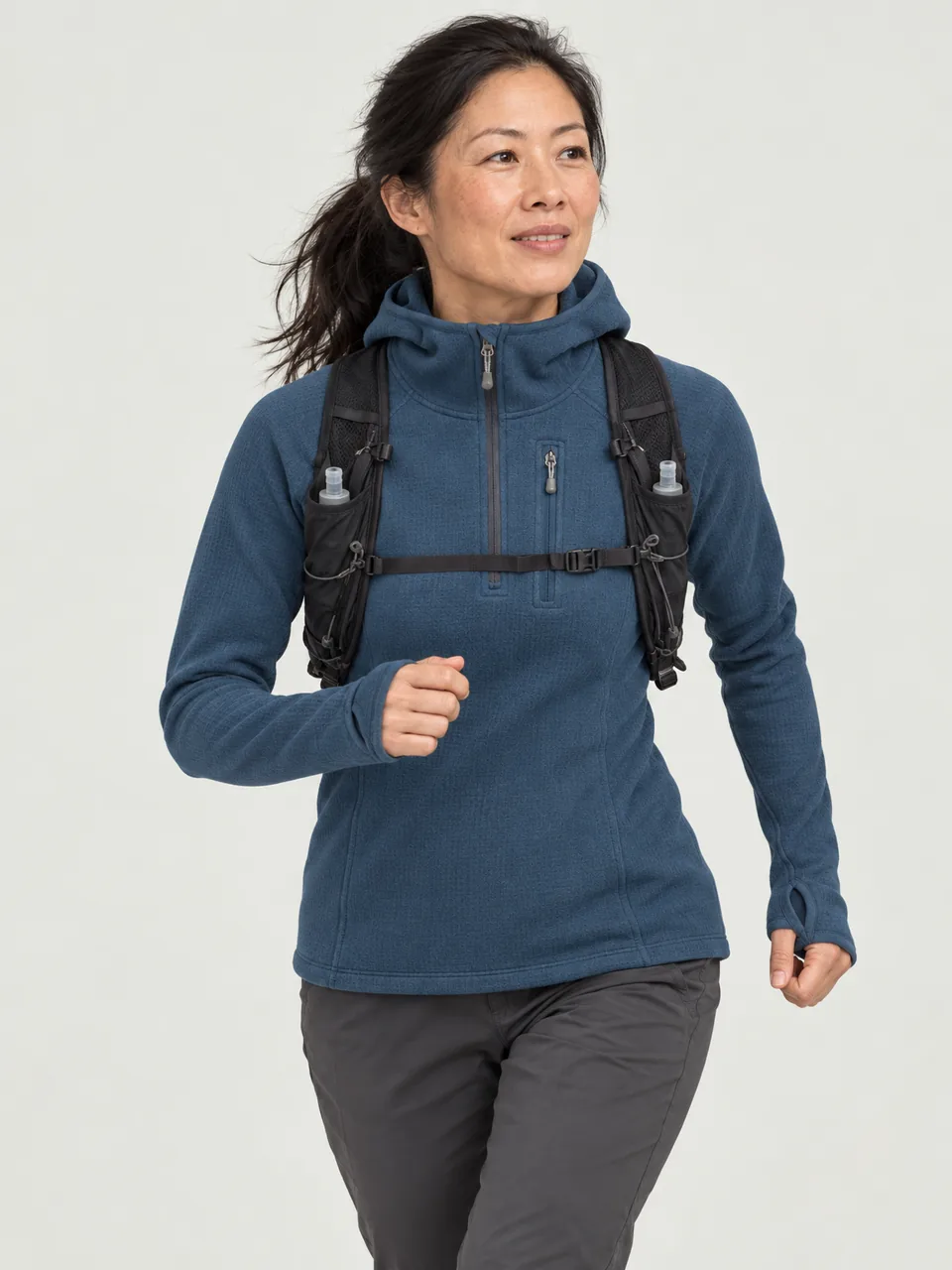 East Asian woman in her 40s, athletic build, jogging down a grassy hillside with a small running vest wearing the Swift Women's Dipper 265 Grid Hoody in Tarn