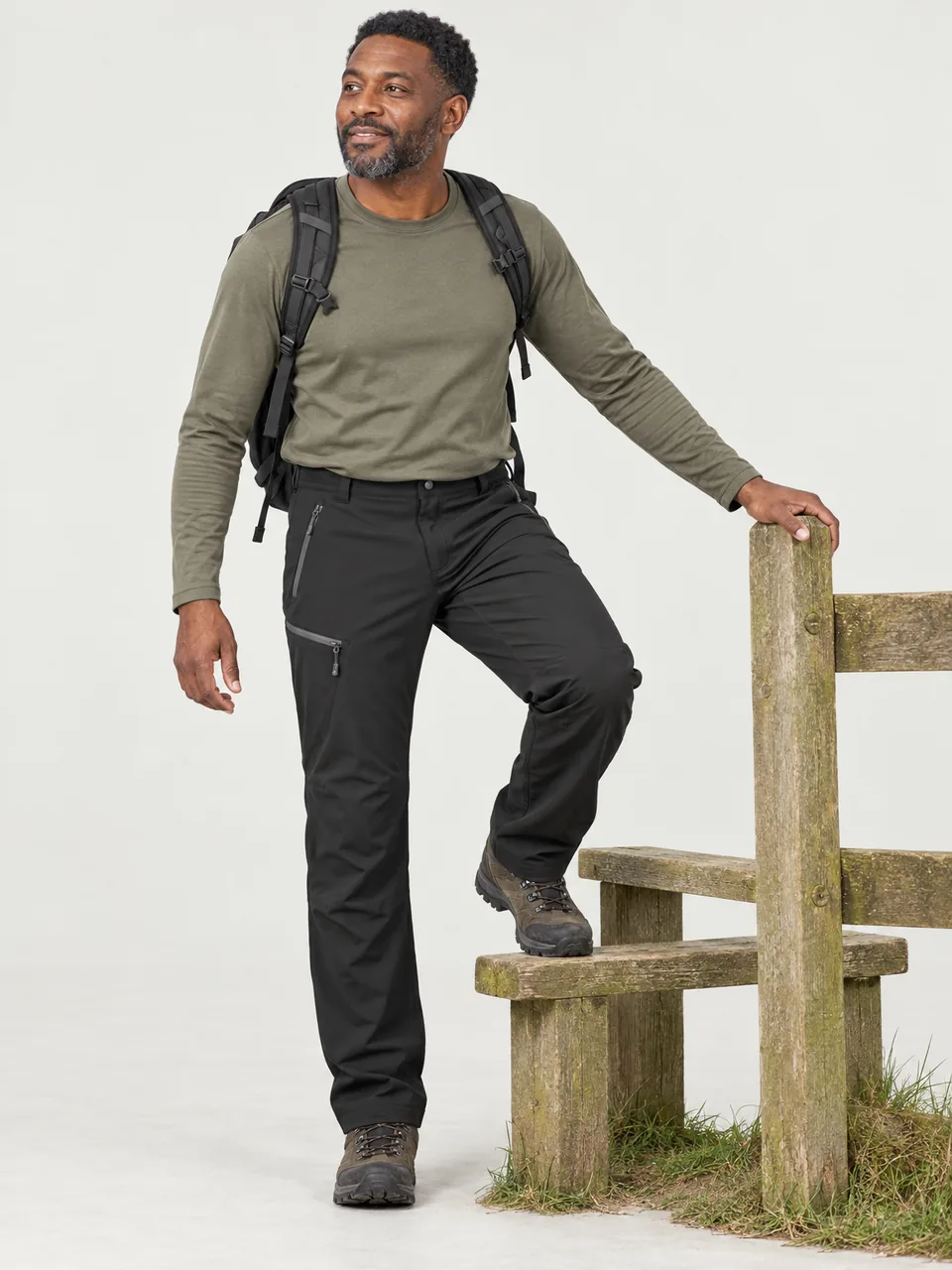 Black British man in his 40s stepping over a stile on a lowland path with a daypack wearing the Hale Men's Inbye Walking Trousers in Soot