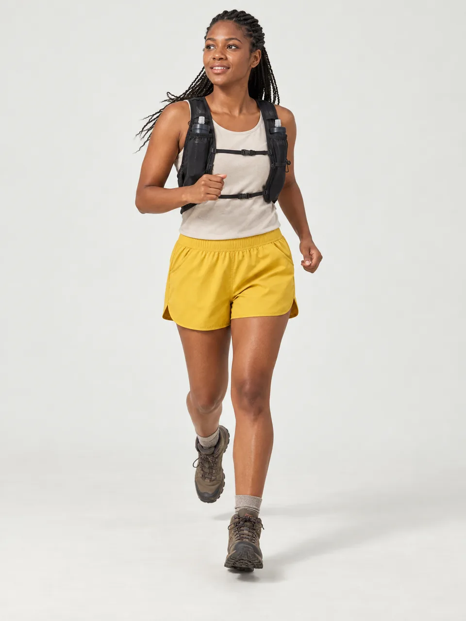 Black woman in her 20s with braided hair, running down a cliff-top path in summer with a small vest pack wearing the Swift Women's Redshank 130 Trail Shorts in Gorse