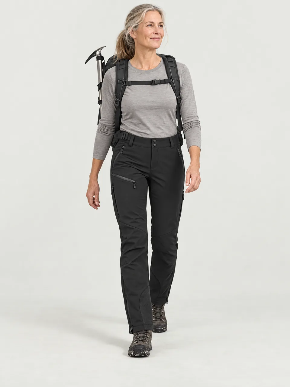 Tall woman in her 50s walking through patchy snow on a high plateau, an ice axe on her pack wearing the Rakefoot Women's Drove Winter Trousers in Black